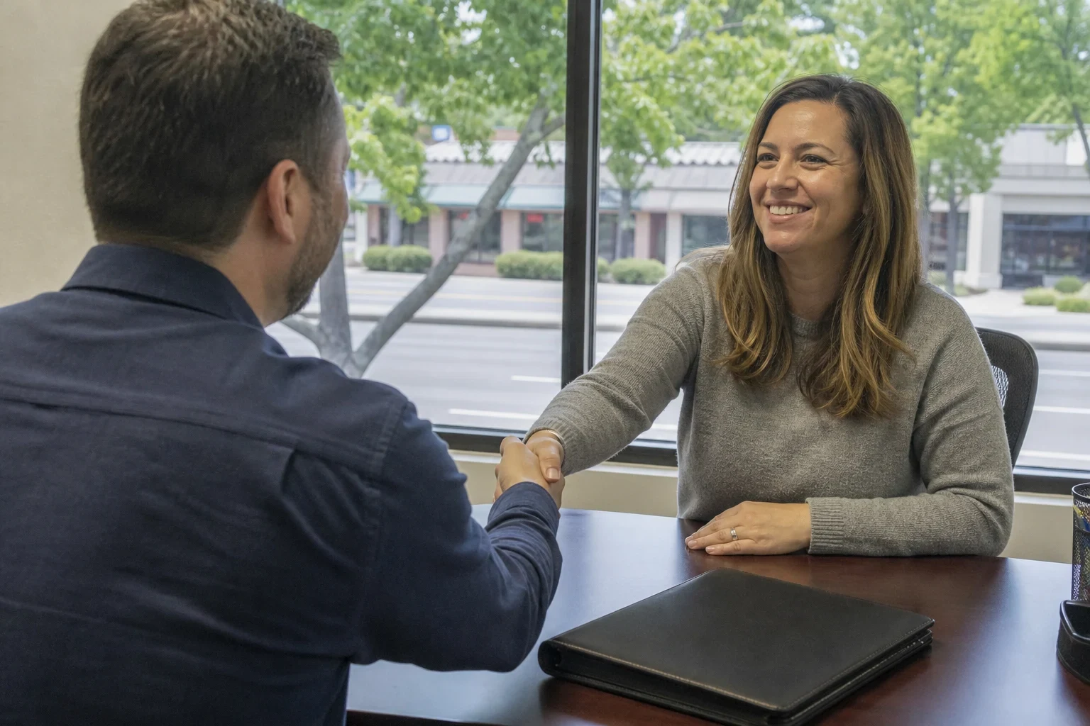 A man in a navy shirt shakes hands with a smiling woman across a wooden desk with a closed black folder, a window showing trees and a strip center behind